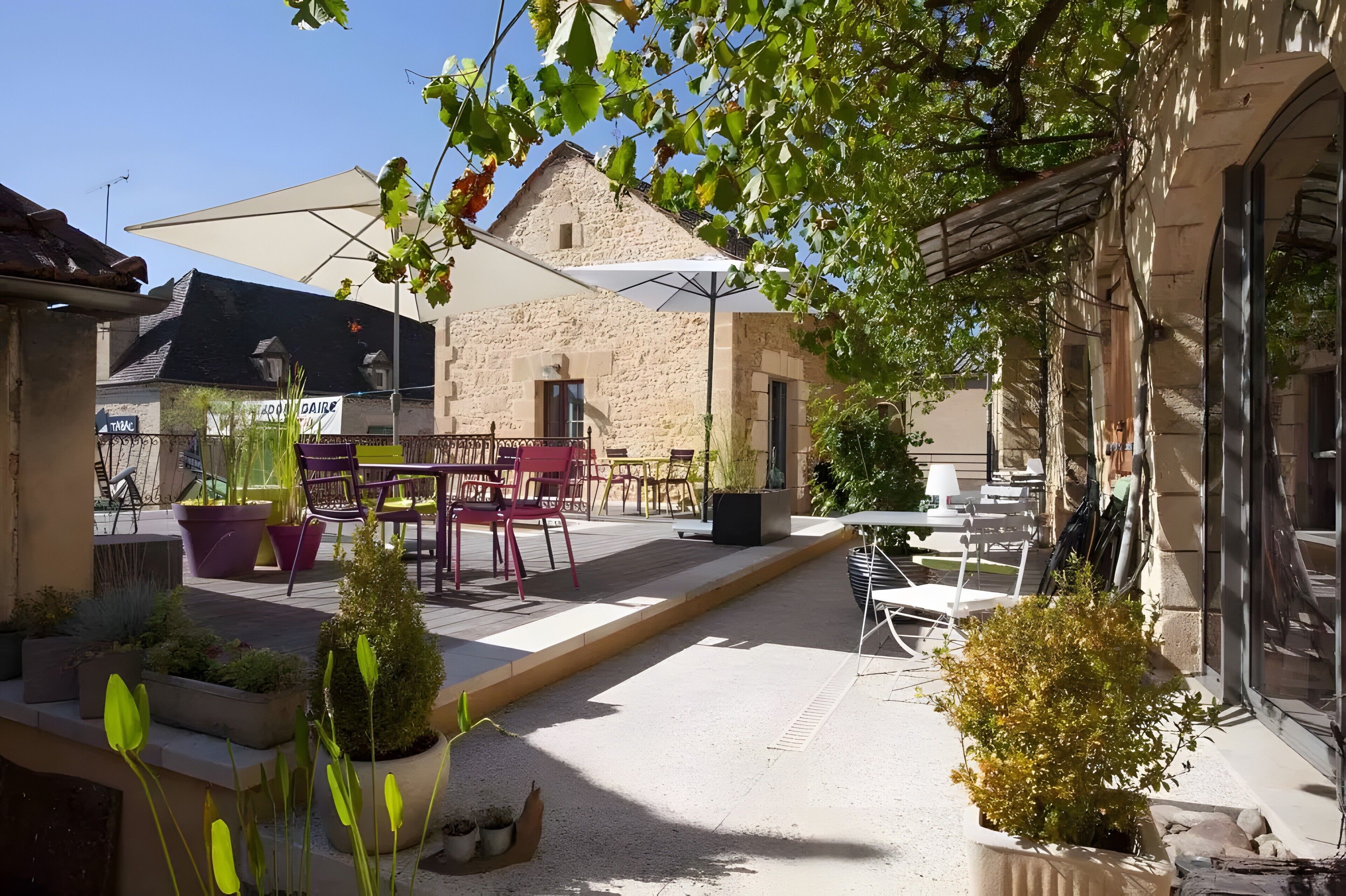 An outdoor café area with tables and chairs, potted plants, and a building with a stone facade.