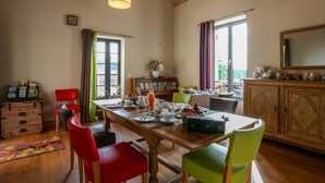 Een eetruimte met een houten tafel, rode en groene stoelen, een kroonluchter en uitzicht op het omliggende landschap door grote ramen.