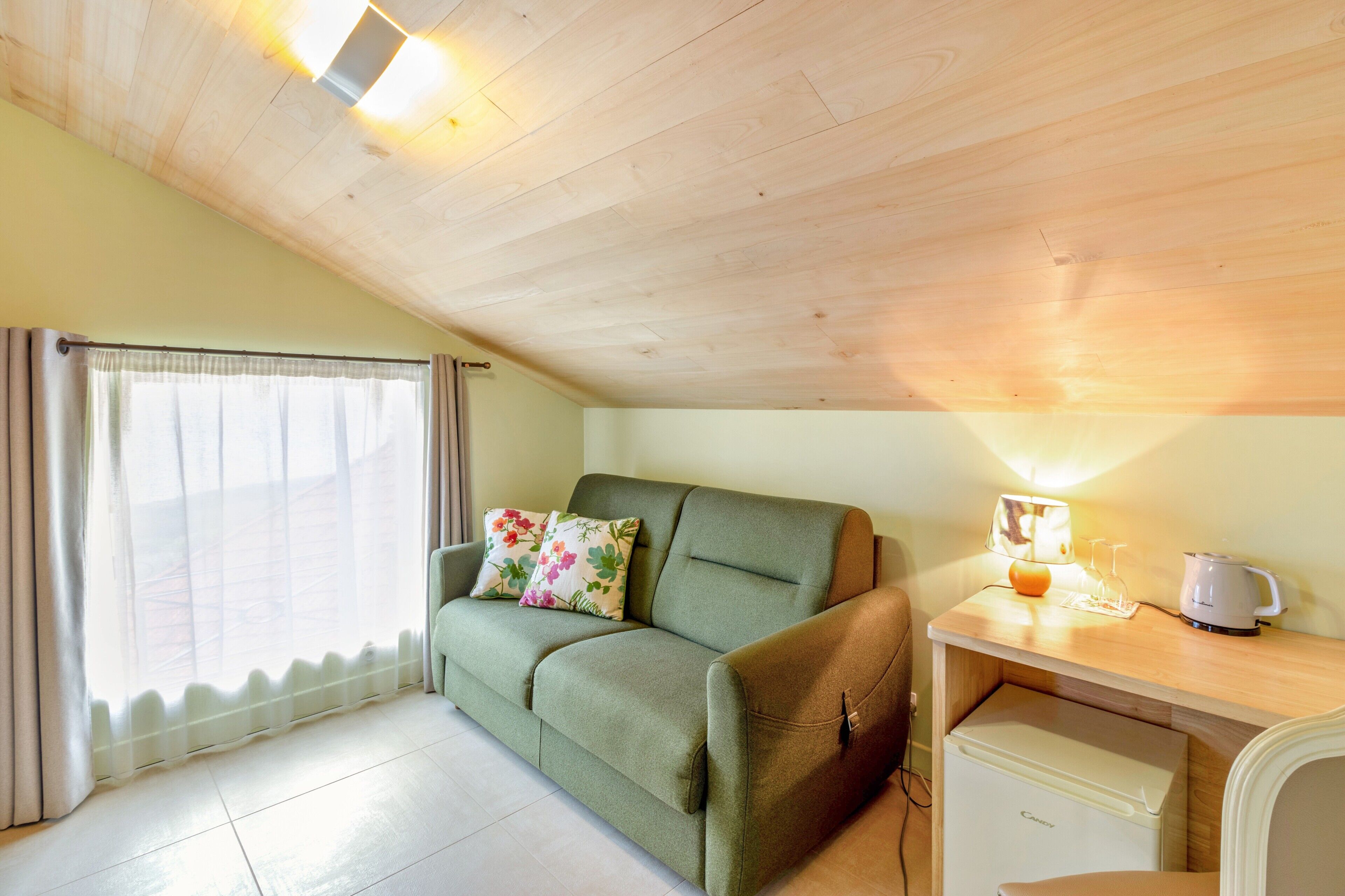 A room with a green sofa, a wooden desk with a kettle, a small refrigerator, and a window with curtains.