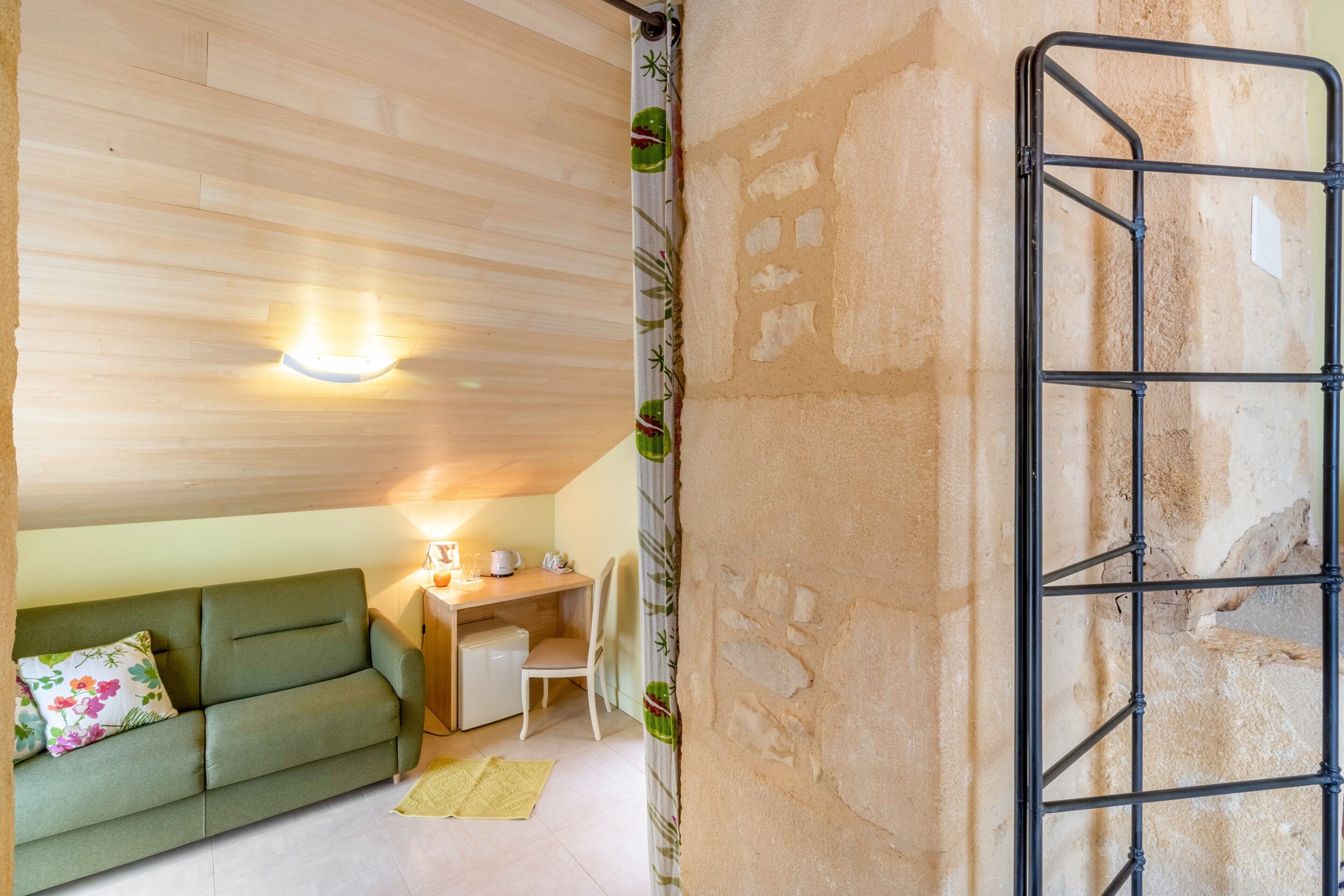 A room with a green sofa, a small desk with a lamp, a chair, and a metal rack against a stone wall.