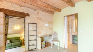 A room with a stone wall, a wooden ceiling, a green sofa, a white chair, a metal shelf, and a bathroom door. - Tilleul Room – Natural Comfort and Spaciousness (Carsac-Aillac)
