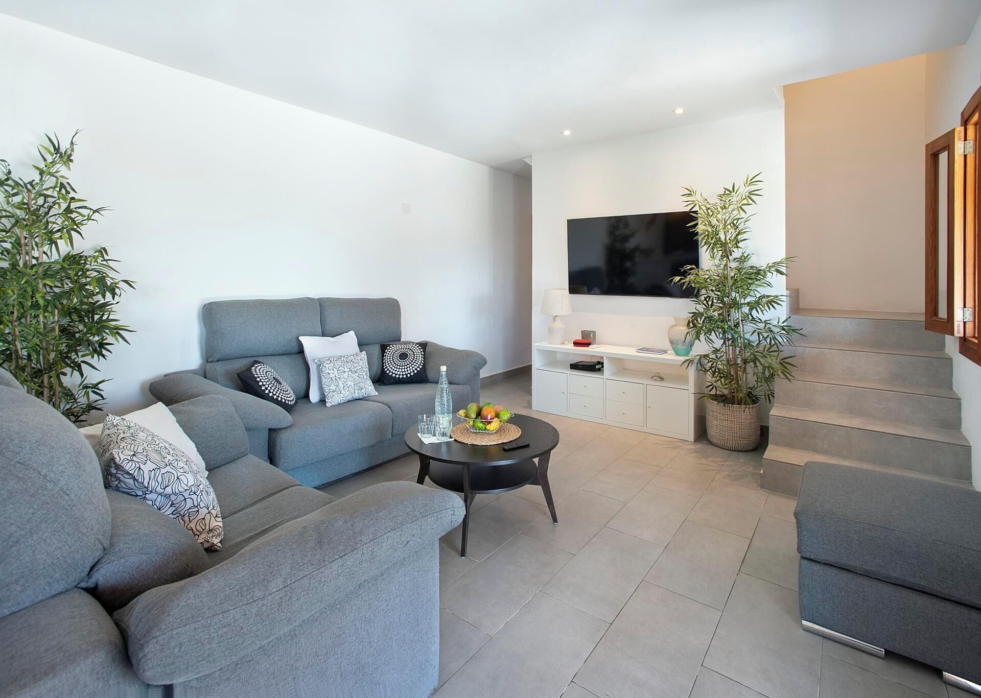A modern living room with grey sofas, a flat-screen TV, a coffee table with fruits, and potted plants.