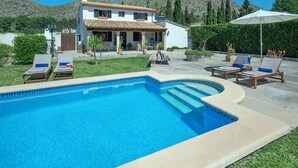 Ein Pool mit Liegestühlen, ein weißes Haus mit Ziegeldach und eine gebirgige Kulisse.