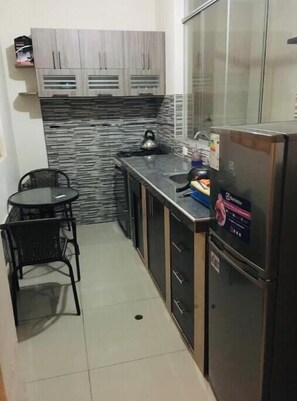 A compact kitchen with grey cabinets, a black countertop, a kettle, a refrigerator, and a small table with chairs.