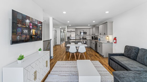 A modern living room with a flat-screen TV, a grey sofa, a dining area with white chairs, and a kitchen in the background. - 3BR Apt Mins to Bus, Newark Airport, Penn Sta, NYC (Newark)