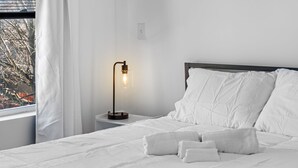 A neatly made bed with white linens and towels, a bedside table with a lamp, and a window with curtains. - 3BR Apt Mins to Bus, Newark Airport, Penn Sta, NYC (Newark)