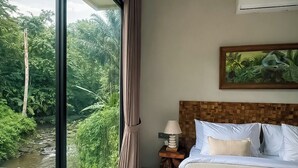 A bedroom with a large window overlooking a river and greenery, a bed with white linens, a wooden headboard, a nightstand with a lamp, and a painting on the wall. - 3-bedroom villa in fantastic ubud Bali with cool AC breeze (Bali)