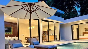 A modern outdoor pool area with lounge chairs and an umbrella, surrounded by glass walls and a wooden deck. - 3-bedroom villa in fantastic ubud Bali with cool AC breeze (Bali)