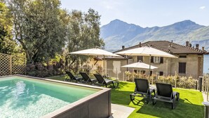 Una piscina con sillas de descanso y sombrillas, un césped cuidado y un edificio con montañas al fondo.