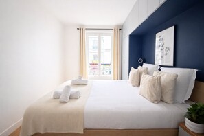 A neatly made bed with white linens and pillows, a bedside plant, and a view of a balcony with outdoor furniture. - Appartement au cœur de Montmartre (Paris)