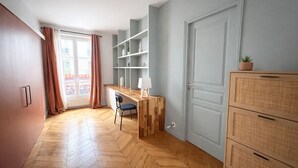 Une pièce dotée d’un parquet en bois à motifs en chevrons, d’un bureau avec une chaise, d’une lampe blanche et d’un meuble en osier.