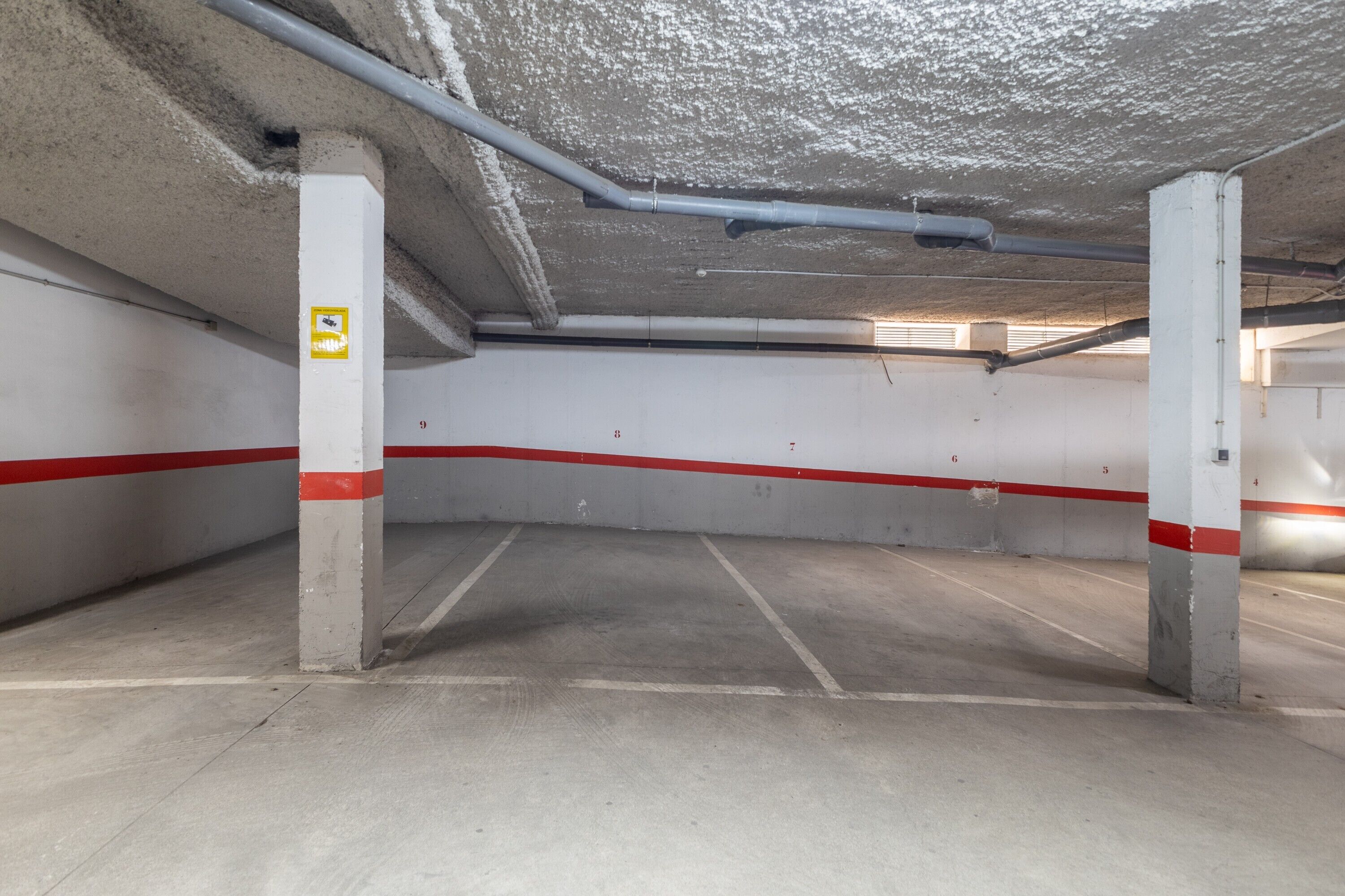 Un parking souterrain avec des piliers en béton, des murs peints en rouge et en blanc, et des tuyaux apparents au plafond.