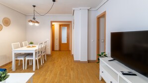 Un salon moderne doté d’un téléviseur à écran plat, d’un coin repas avec une table et des chaises blanches, et d’un couloir menant à une autre pièce.