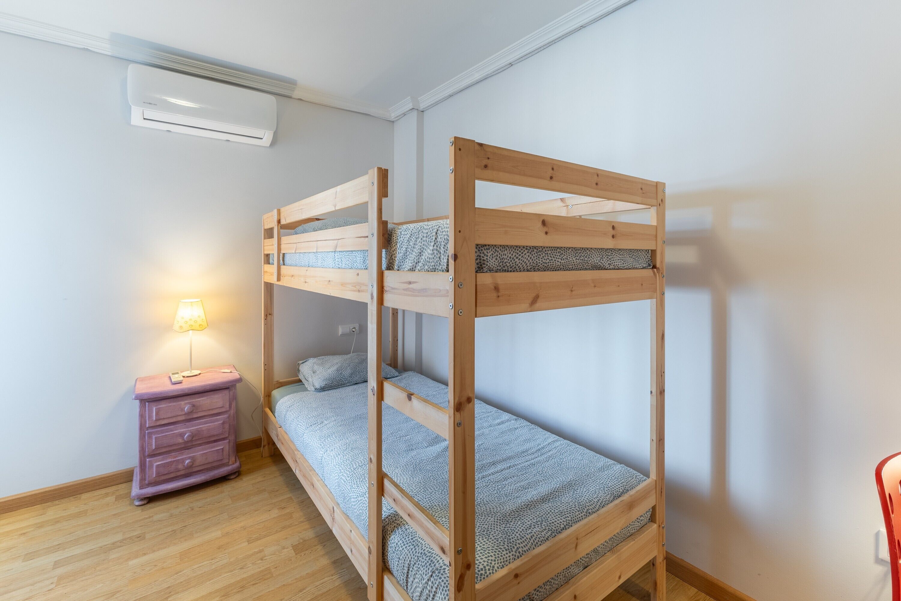 Una cama litera con estructura de madera y ropa de cama estampada, una mesita de noche pequeña con lámpara, y un aire acondicionado instalado en la pared.