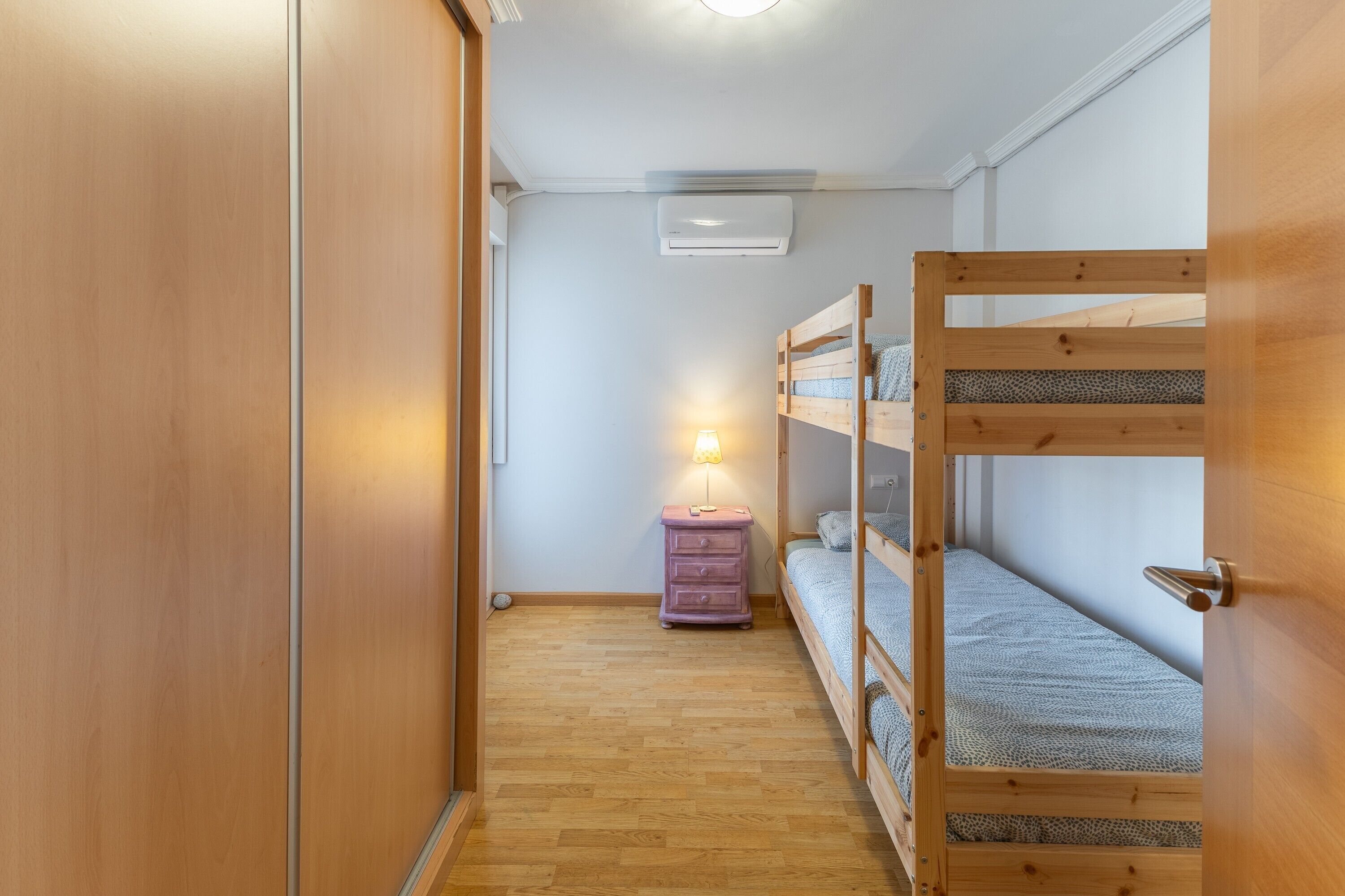 Un couloir étroit avec un plancher en bois, un lit superposé, une petite commode avec une lampe et un appareil de climatisation fixé au mur.
