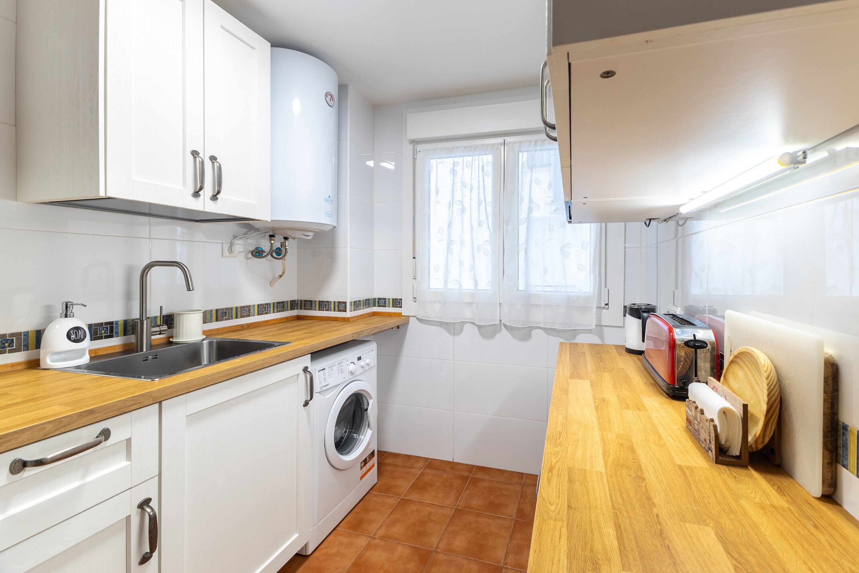 Een kleine keuken met witte kasten, een wasmachine, een spoelbak en een houten aanrecht.