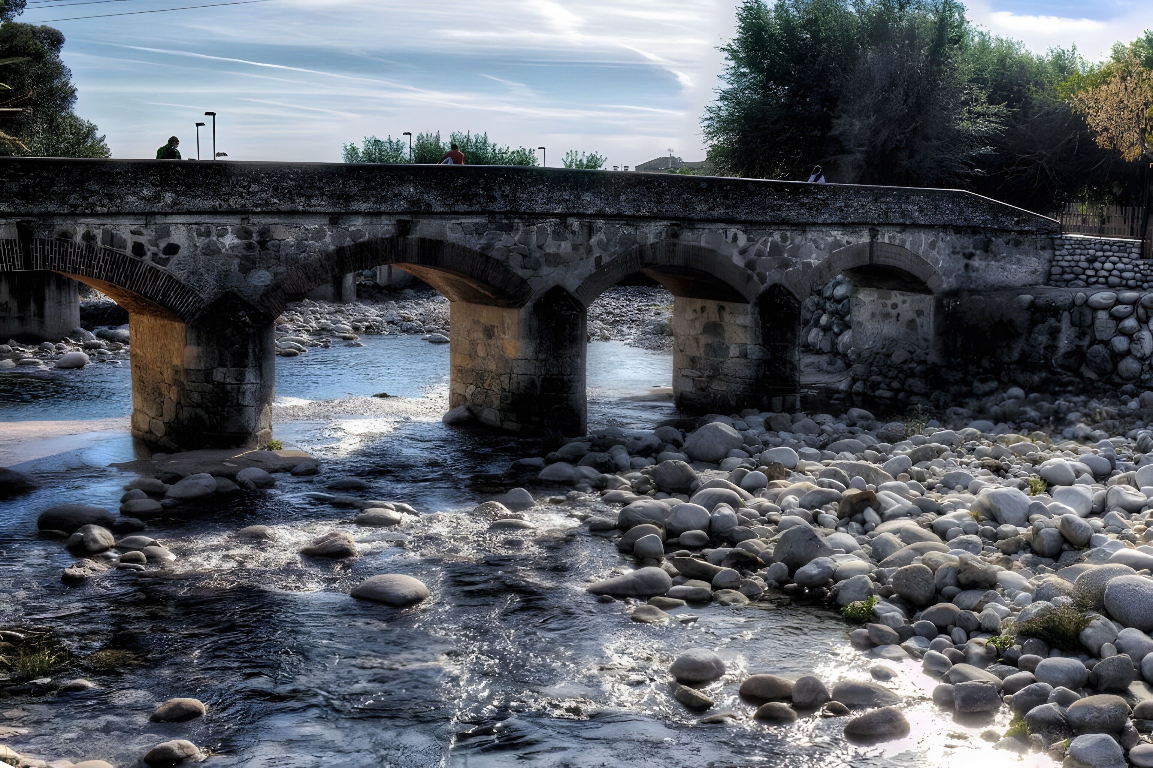 A stone bridge over a river with a rocky bed, surrounded by trees and a clear sky.