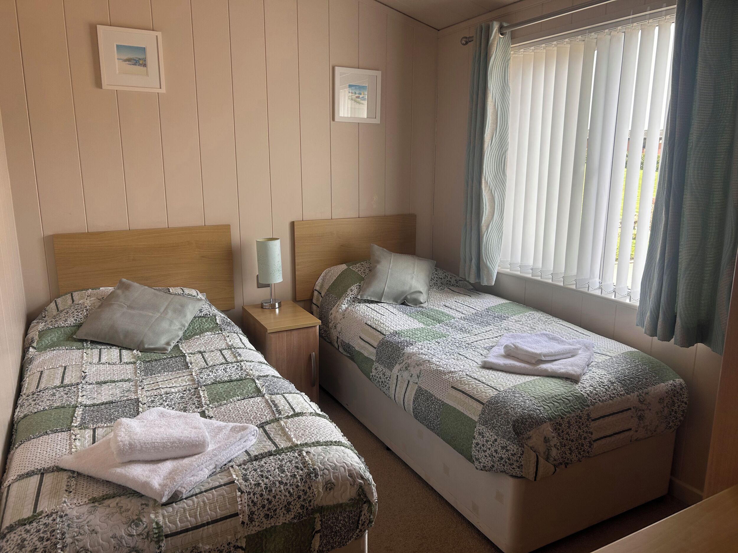 Quarto com duas camas, uma mesa de cabeceira com abajur e uma janela com cortinas.
