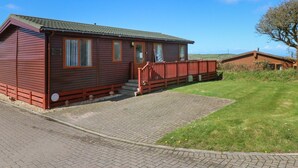A single-story wooden cabin with a porch, a brick driveway, and a grassy yard. - 1 Ocean Terrace (Ilfracombe)