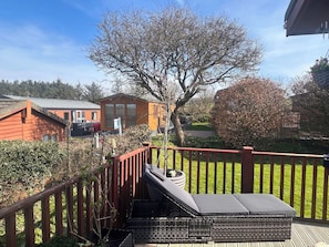 En terrass med en flätad loungestol, ett träräcke och utsikt över en röd byggnad och ett träd.