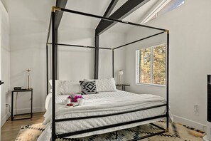 Una cama con dosel en una habitación con un ventanal grande, una mesita de noche pequeña y una alfombra.