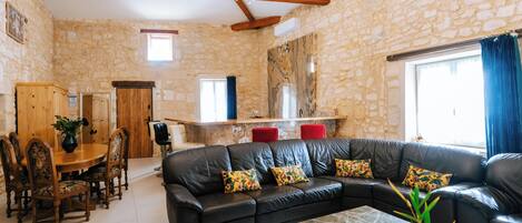 A spacious living area with a black leather sofa, wooden dining table, and chairs, stone walls, and a chandelier.