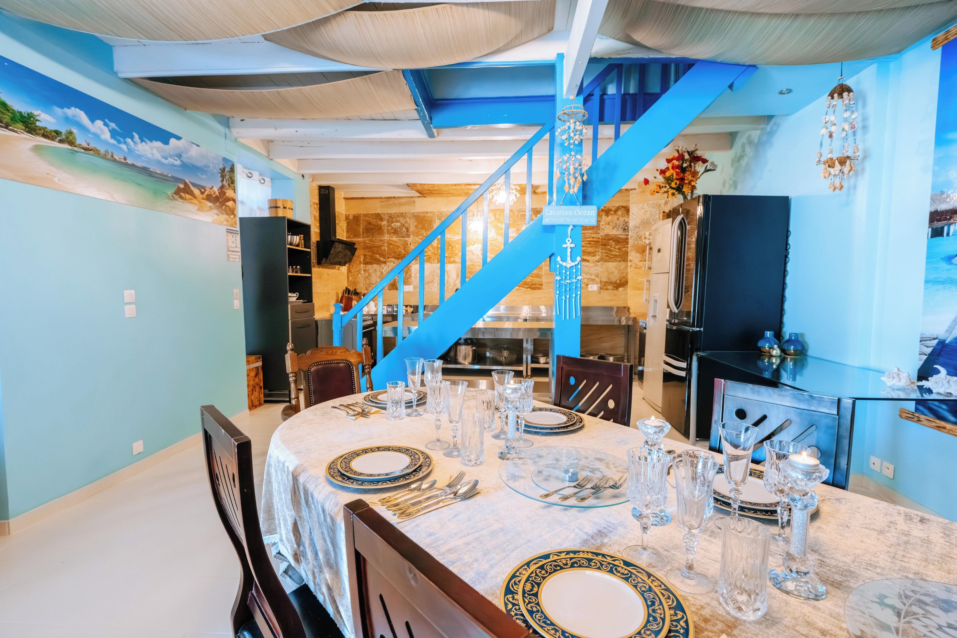 Un coin repas avec un escalier bleu, une fresque représentant une plage et une cuisine en arrière-plan.