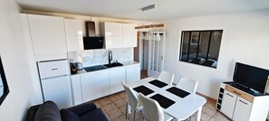 A compact kitchen with white cabinets, a built-in oven, and a dining area with a white table and chairs. - New: 1-Bedroom Apartment Facing the Sea, Balcony, Wi-Fi, Secure Garage, Near Town Center (Saint-Vaast-la-Hougue)