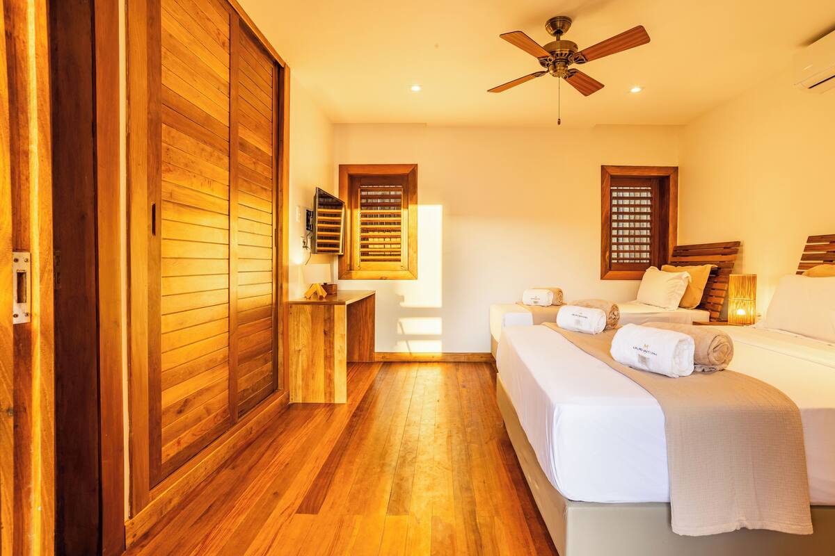 Une chambre avec un plancher en bois, un lit recouvert de draps blancs, une armoire en bois et un ventilateur de plafond.
