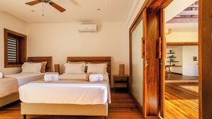 A hotel room with a large bed, two smaller beds, wooden flooring, a ceiling fan, and an air conditioning unit.