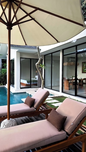 Un moderno espacio lounge al aire libre con dos sillones acolchados y una sombrilla grande, con vistas a una piscina y un área de comedor con paredes de vidrio.