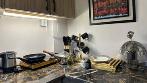 A kitchen with a stove, frying pan, kettle, and various utensils. There is a framed picture on the wall and a countertop with a plate and salt and pepper shakers.
