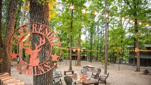Un'area salotto esterna rustica con un braciere, circondata da alberi e luci decorative.