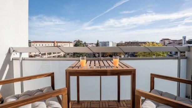 Un balcón con una mesa de madera y dos sillas, dos vasos de jugo de naranja, y vistas a edificios al fondo.