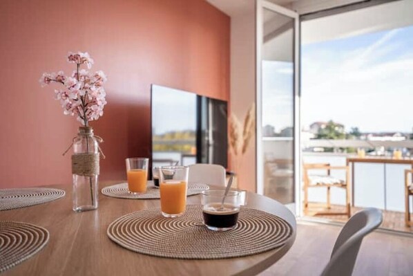A dining table with two glasses of orange juice and a bottle of water, a vase with pink flowers, and a television in the background.