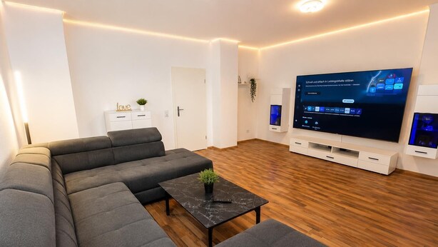 A modern living room with a sectional sofa, a black coffee table, a flat-screen TV, and a wooden floor. - 70m² suite incl. 98 "Smat-TV + parking lot + balcony (Chemnitz)