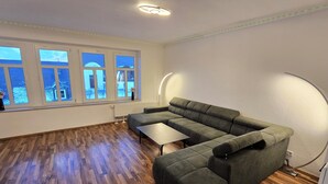 A modern living room with a sectional sofa, a small table, and large windows with a view of buildings. - 70m² suite incl. 98 "Smat-TV + parking lot + balcony (Chemnitz)