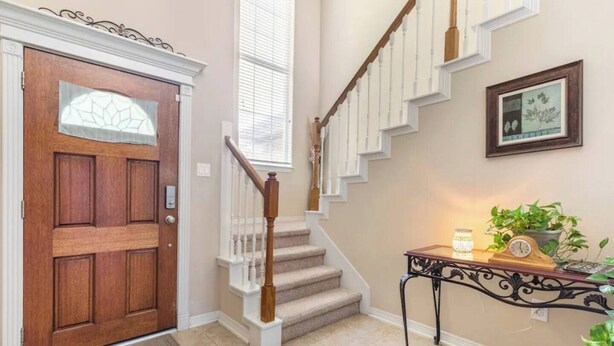 A wooden front door with a glass panel, a staircase with white railings, and a small table with a clock and plants.