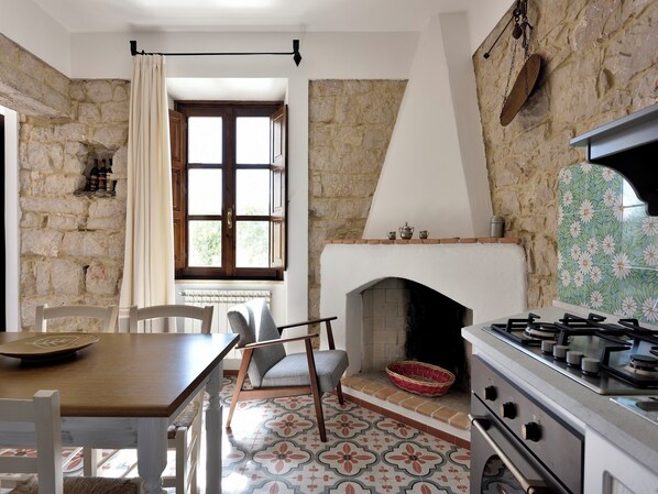 A rustic kitchen with a stone wall, a fireplace, a dining table with chairs, and a window with curtains. - Sea and mountain views near Cala Goloritze (Baunei)