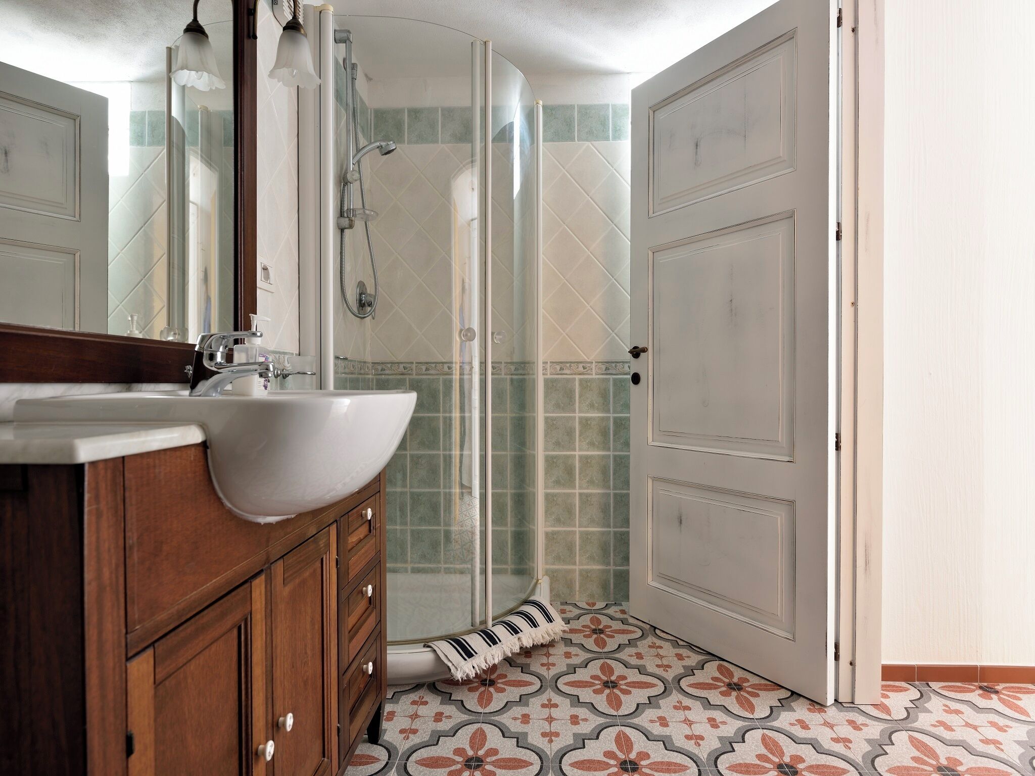 Un baño con un lavamanos blanco, tocador de madera, cabina de ducha con mampara de cristal y baldosas de suelo estampadas.