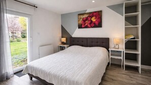 A bedroom with a large bed, a nightstand with a lamp, a wardrobe, and a window with a view of a garden. - Gîte Le Laurier (Villebernier)