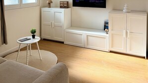Una sala moderna con un sofá, una mesita redonda con una planta, un televisor en la pared y armarios blancos.