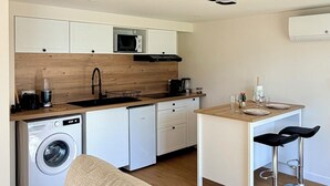 Una cocina compacta equipada con lavadora, microondas y cafetera. Cuenta con un área de comedor con dos taburetes altos y una mesa pequeña.