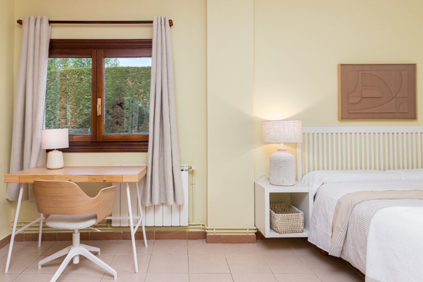 Un dormitorio con cama, mesita de noche, lámpara, silla, escritorio, ventana con cortinas, radiador y una obra de arte colgada en la pared.
