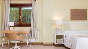 Un dormitorio con cama, mesita de noche, lámpara, silla, escritorio, ventana con cortinas, radiador y una obra de arte colgada en la pared.