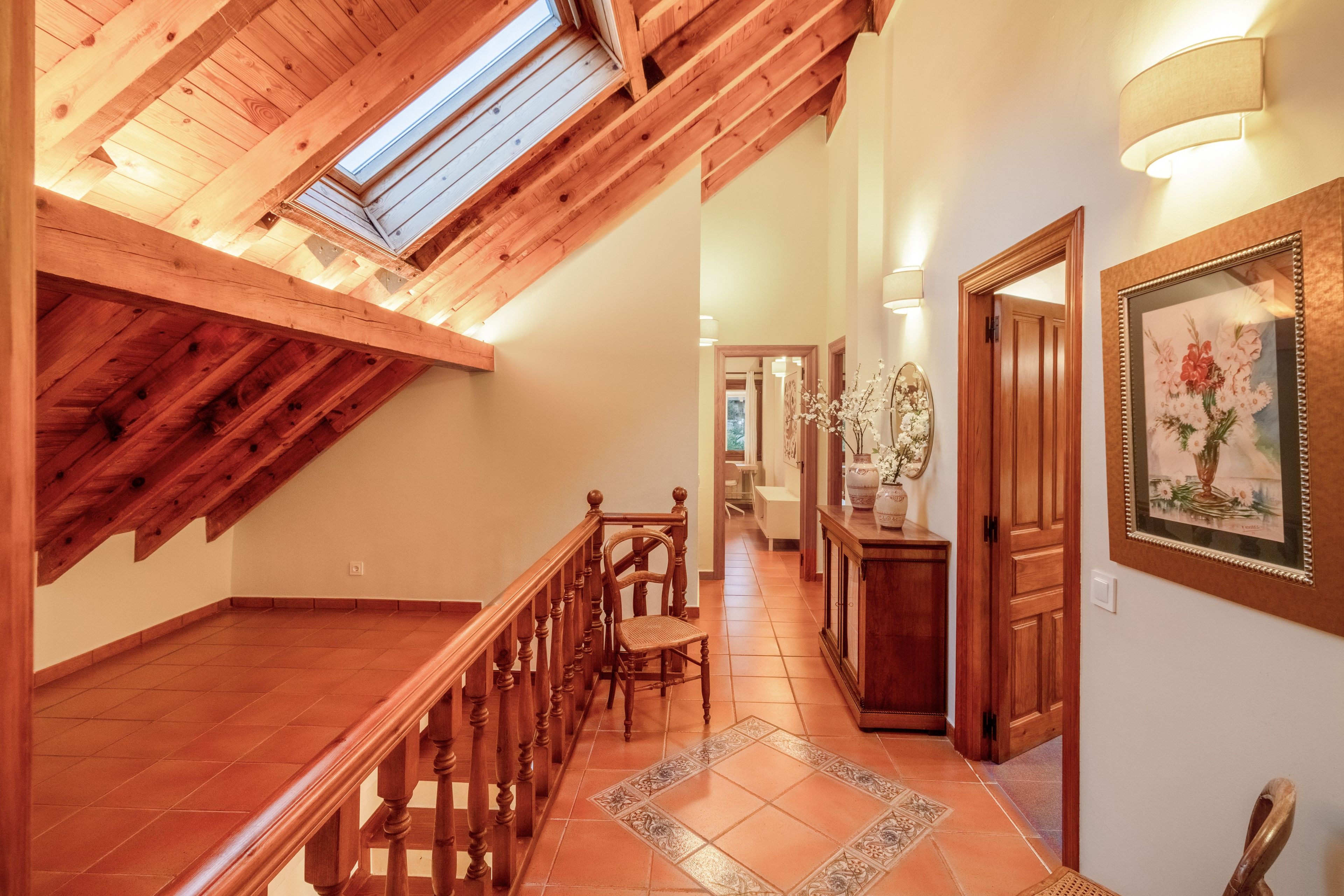 Una escalera de madera que conduce a un nivel superior con un tragaluz, un pasillo con una puerta y un cuadro enmarcado, y un piso de baldosas.