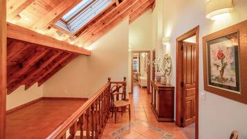 Una escalera de madera que conduce a un nivel superior con un tragaluz, un pasillo con una puerta y un cuadro enmarcado, y un piso de baldosas.