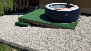 Un jacuzzi sobre un tapete verde, una terraza de madera y un edificio color crema con un cielo azul claro al fondo.