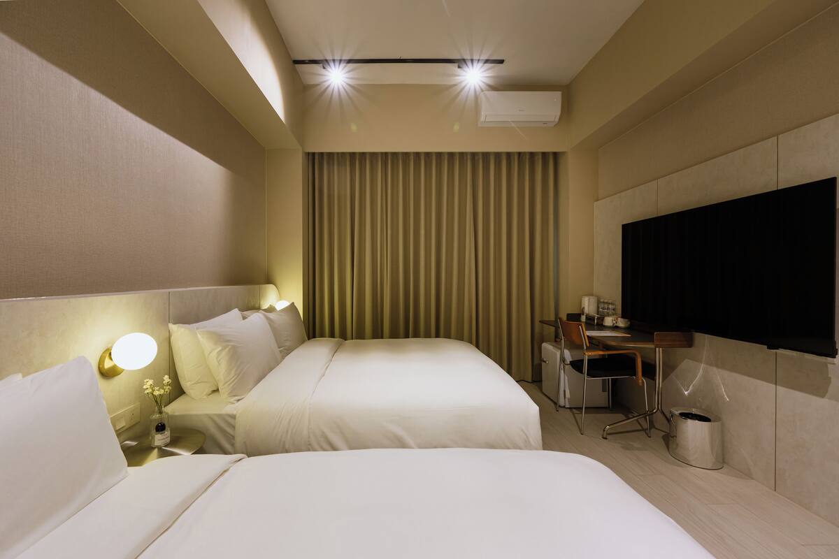 A hotel room with two beds, a desk, a chair, a TV, and a curtain.