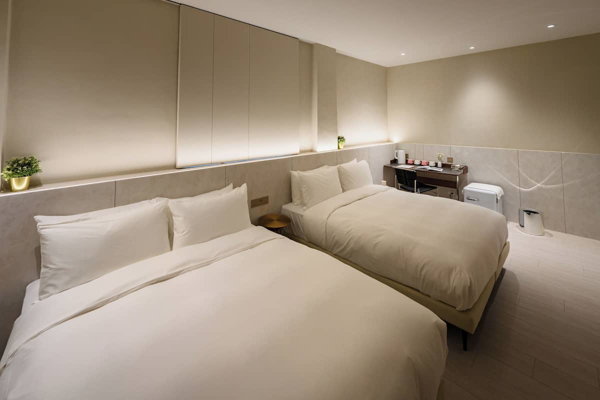A modern hotel room with two beds, a bedside table with a lamp, a small plant, and a trash bin.